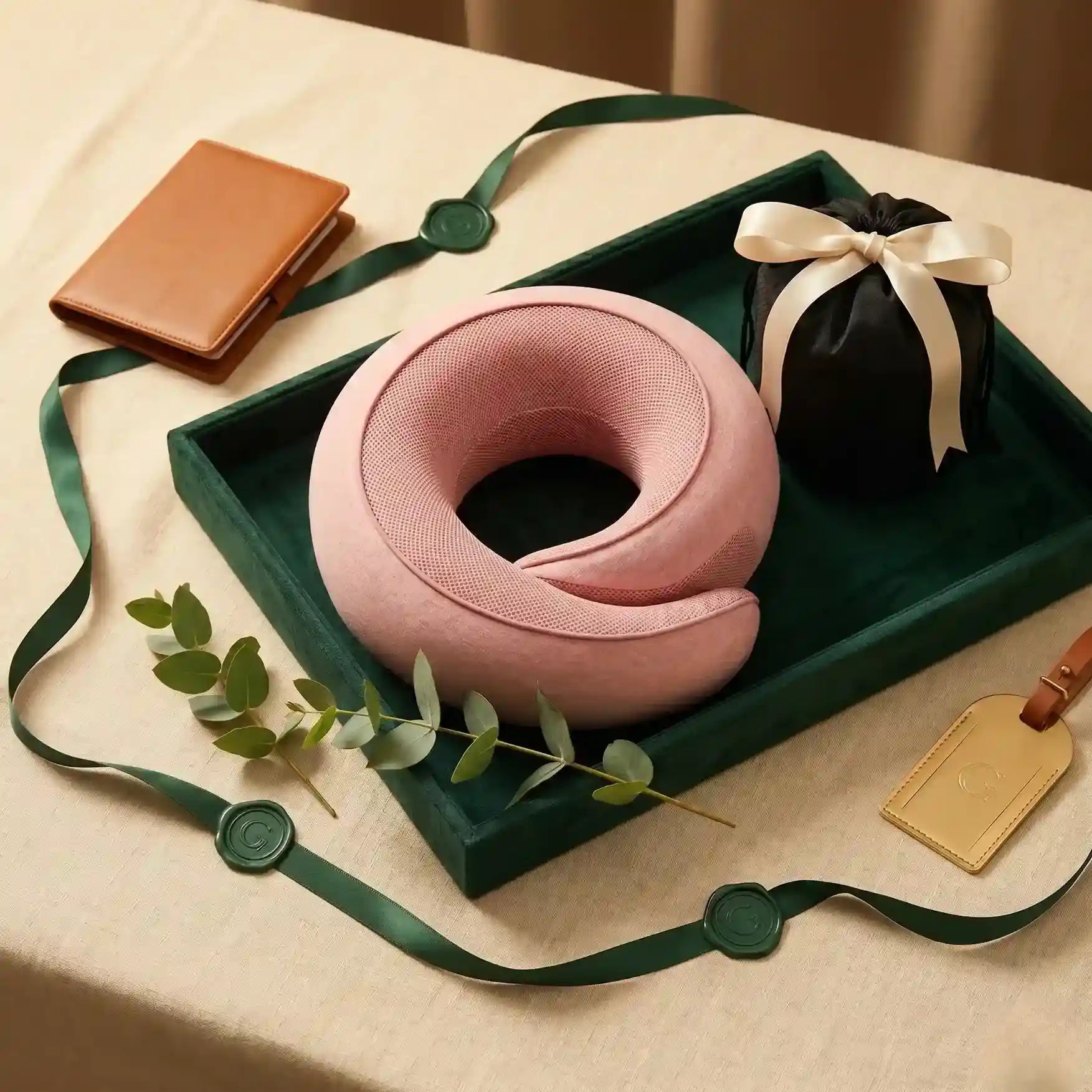 Pink neck pillow on a green tray with a brown leather case and black pouch on a beige surface.