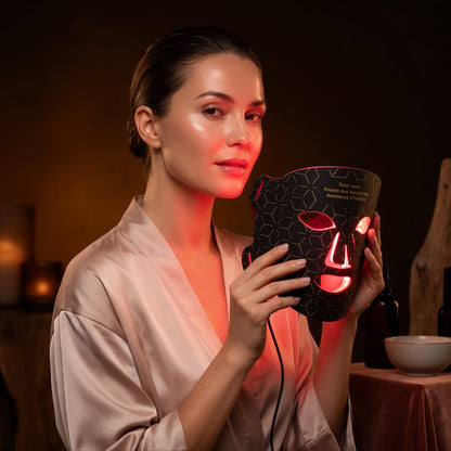 A woman in a satin robe holds a black LED facial therapy mask with red light glowing from the cutouts, set against a dimly lit spa-like background.